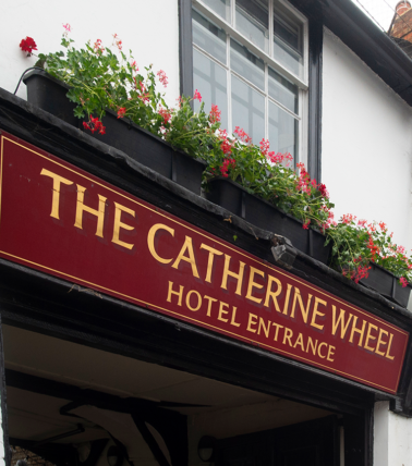 Exterior sign of The Catherine Wheel hotel entrance sign with red flowers in planters above